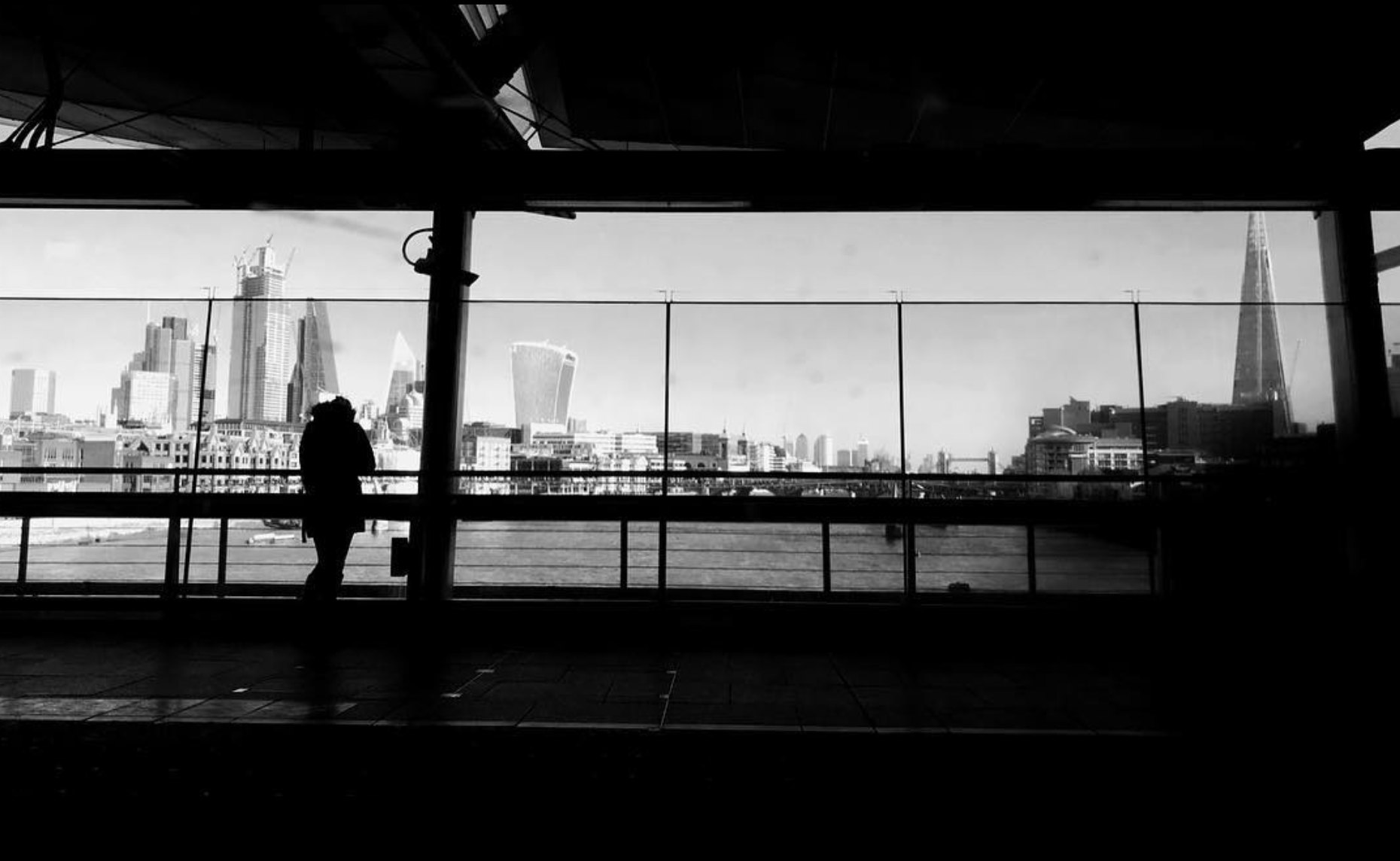 A figure silhouetted against the City of London skyline, viewed through a riverside walkway.